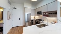 This thoughtfully designed single-floor residence blends contemporary style with warm, curated details throughout. The open-concept kitchen is outfitted with light wood cabinetry, quartz countertops, stainless steel appliances including a French door refrigerator, a wine cooler, and a gas range, along with open shelving for a modern, functional aesthetic. Hardwood flooring flows seamlessly through the space, connecting each room with a cohesive warmth.

The living area is a standout, anchored by a textured blue-grey wallpapered accent wall, a blush velvet sofa, and a striking globe pendant light. A round gold-toned mirror adds a reflective, decorative focal point. Floor-to-ceiling windows and a glass door open to a private balcony, offering views of the surrounding cityscape and filling the room with abundant natural light.

The bedroom continues the refined design language with the same textured wallpaper, a dramatic chain-link chandelier, and a walk-in closet with built-in shelving. Curated artwork adorns the walls, adding personality and depth to the space.

The bathroom features soft blush-toned walls accented by a thoughtful gallery of framed artwork, creating a calm and welcoming atmosphere. Every corner of this residence reflects intentional design and careful attention to detail.