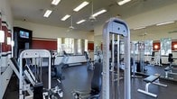 This fitness center offers a comprehensive workout environment outfitted with professional-grade equipment from Inflight Fitness. The space is generously sized, providing ample room between machines for comfortable, unrestricted movement. Strength training is well-represented with cable machines, weight stacks, and resistance equipment positioned throughout the floor. Cardio options, including treadmills and other aerobic machines, are arranged along the perimeter near the windows. Large mirrors line the walls, enhancing the sense of openness and allowing users to monitor their form during exercise. The flooring consists of durable, dark rubber matting suited for heavy equipment and high-traffic use. Elevated ceilings fitted with ceiling fans and recessed fluorescent lighting contribute to a well-ventilated, evenly lit atmosphere. Natural light filters in through a row of windows along one wall, offering views of the surrounding greenery and adding an inviting quality to the space. Accent walls in deep red add a bold visual contrast to the otherwise neutral color palette. The overall design prioritizes functionality and accessibility, making it a practical and well-appointed facility for a variety of fitness routines.