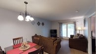 This single-floor residence offers a welcoming and functional layout centered around an open-concept living and dining area. The dining space is anchored by a classic chandelier with bell-shaped shades, casting warm light over a wood dining table set with red placemats and surrounded by sturdy wooden chairs. The living area flows seamlessly from the dining space, furnished with plush brown sofas arranged around a flat-screen television and a cozy fireplace, creating an inviting atmosphere for relaxation and entertainment. Large sliding glass doors along the far wall flood the room with generous natural light and offer a view of the lush greenery outside, enhanced by patterned curtains that add a decorative touch. The walls are finished in a soft, neutral blue-gray tone, complementing the warm brown tones of the furniture and carpeted flooring throughout. A textured ceiling adds subtle architectural character to the space. Decorative diamond-shaped wall accents provide visual interest without overwhelming the clean, understated aesthetic. Overall, the space balances practicality with comfort, making it an ideal everyday living environment.
