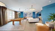 Located at 4400 Calvert Rd in College Park, Maryland, this single-floor amenity lounge is a thoughtfully designed social and recreation space that combines modern aesthetics with a lively, welcoming atmosphere. The room is defined by striking cobalt blue accent walls that create a bold visual contrast against the neutral gray tile flooring and white ceiling, which is adorned with a series of large, elegant drum pendant lights that illuminate the space warmly.

A dramatic large-scale marbled mural in swirling shades of blue and white commands attention as a central focal point, anchoring the entertainment zone. This area is equipped with a full-size billiards table and a shuffleboard table, offering ample recreational options. Tall windows dressed in warm linen-toned curtains allow generous natural light to pour in, enhancing the open, airy feel of the room.

Multiple seating vignettes are distributed throughout, including plush white sofas arranged on a patterned area rug, and intimate conversation nooks featuring curved cognac leather chairs and round marble-topped accent tables. Tropical potted plants are placed throughout, adding organic texture and a refreshing natural element. A built-in bar or kitchenette area is visible at the rear, completing the space as an all-inclusive social hub ideal for gatherings and leisure.