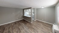 This single-floor unit offers a practical and functional layout connecting a living area, kitchen, bedroom, and bathroom in a compact, efficient floor plan. The living area is finished with wood-look plank flooring in a warm brown tone, complemented by neutral gray walls and a textured ceiling. A wall-mounted air conditioning unit and baseboard heating provide year-round climate control, while a window admits natural light. The kitchen is accessible via a pass-through opening with a breakfast bar counter, creating an open connection to the living space. White appliances, including a range and refrigerator, are visible within the kitchen, which also features white cabinetry. The carpeted bedroom is finished in the same neutral gray palette and is equipped with a baseboard heater and a window overlooking an exterior parking area. The bathroom features white ceramic tile lining the walls of a combination bathtub and shower enclosure, with a small window positioned above the tub to provide ventilation and natural light. Throughout the unit, clean white baseboards and consistent neutral tones create a cohesive, understated aesthetic.