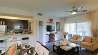 This single-floor residence showcases an open-concept design that integrates the living, dining, and kitchen areas into one cohesive space. The living area is anchored by a ceiling fan with an integrated light fixture, providing both comfort and illumination. A sliding glass door floods the space with natural light and offers access to an outdoor area, enhancing the overall sense of openness. The entertainment setup features a flat-screen television mounted on a distinctively tiered wooden media stand, adding a sculptural focal point to the room. Hardwood-style flooring runs throughout, lending warmth and continuity across the space. The dining area accommodates a glass-top table with dark chairs, while the kitchen, visible through a pass-through opening, features dark cabinetry, a tile backsplash, and a breakfast bar with seating. Decorative accents, including wall art and floral arrangements, contribute to the space's polished, contemporary aesthetic. The neutral white walls and clean architectural lines create a bright, welcoming ambiance throughout.