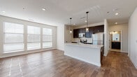 This well-appointed single-floor residence offers a thoughtfully designed open-concept layout that seamlessly connects the kitchen, dining, and living areas. The kitchen is a standout feature, showcasing dark espresso cabinetry, a white quartz waterfall island with an integrated sink, subway tile backsplash, and a full suite of stainless steel appliances including a range, over-the-range microwave, and refrigerator. Pendant lighting above the island adds a modern touch, while recessed ceiling lights illuminate the space evenly. Rich hardwood-style flooring flows throughout the main living areas, complemented by large windows with blinds that invite generous natural light and offer views of the surrounding neighborhood. The living area is spacious and open, with clean white walls that create a bright, airy ambiance. The bedrooms are carpeted for comfort, each featuring large windows and ample closet space. One bedroom includes two separate doorways, suggesting additional storage or a walk-in configuration. The two bathrooms are finished with light tile flooring and dark vanities; one features a framed glass shower enclosure, while the other includes a full soaking tub with a shower combo, both appointed with brushed bronze fixtures.