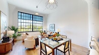 This single-floor residence showcases a thoughtfully designed open-concept layout that blends industrial character with contemporary refinement. The living and dining area is anchored by a dramatic floor-to-ceiling factory-style window with bold black steel framing, flooding the space with natural light and offering expansive views of the surrounding treetops. Soaring ceilings amplify the sense of openness, while geometric pendant light fixtures add visual interest overhead. Light-toned hardwood flooring runs throughout, unifying each area of the home seamlessly.

The kitchen is a standout feature, centered around a generously sized waterfall island topped with veined stone countertops in rich, dark tones. Two-tone cabinetry — combining white uppers with dark lower cabinets — is complemented by a geometric tile backsplash and stainless steel appliances, creating a refined, modern aesthetic. A cluster pendant light fixture above the island adds a sculptural touch.

The bedroom is calm and understated, with soft neutral walls, continued hardwood flooring, and ample wall space for art. A marble-topped dresser and clean architectural details contribute to the room's polished, serene atmosphere. Throughout the unit, the design balances loft-inspired industrial elements with warm, livable finishes.