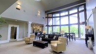 This single-floor space makes a striking first impression with its dramatic vaulted ceilings that rise to a considerable height, creating an expansive, airy atmosphere throughout. A dominant wall of floor-to-ceiling windows with dark framing floods the interior with natural light and offers open views of the surrounding greenery, blurring the boundary between indoors and outdoors.

The main living area is anchored by a dark leather sofa accented with decorative pillows, complemented by cushioned swivel chairs in a warm neutral tone and a tufted leather ottoman serving as a central coffee table. The seating arrangement is thoughtfully positioned to take full advantage of the natural light and outdoor views.

Adjacent to the living area, a dining space features a dark table with matching chairs, positioned near the window wall to benefit from the natural light. Beyond, a kitchen area is visible, defined by a raised bar with seating, pendant lighting, and stainless steel appliances. Decorative columns provide subtle architectural definition between zones. The overall ambiance is modern, open, and welcoming, with a cohesive neutral palette throughout.