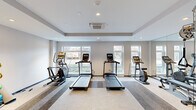 This fitness center is a thoughtfully designed single-floor space built to support a comprehensive workout experience. The room features a clean, modern aesthetic with neutral gray walls, a tray ceiling with recessed lighting, and a cassette-style HVAC unit for climate control. Large windows line the walls, flooding the space with natural light and offering views of the surrounding outdoor greenery.

The cardio zone is well-appointed with multiple treadmills, an elliptical trainer, a stair climber, and a stationary bike, all arranged to make the most of the natural light from the windows. A full-length mirror wall visually expands the space and allows users to monitor their form during exercise.

The strength and functional training area includes a multi-functional cable machine and a rack of medicine balls and exercise balls in various sizes, providing options for a wide range of workouts. A flat workout bench adds to the versatility of the strength training zone.

A dedicated stretch and open-floor area offers ample room for floor exercises, yoga, or cooldown routines. Practical amenities include a dual-level water fountain and a changing room with a shower, ensuring convenience for users before and after their sessions. The patterned carpet flooring with striped detailing adds a polished, professional finish to the overall space.