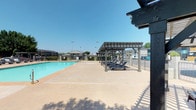 Located in Abilene, this outdoor amenity space centers around a generously sized swimming pool with clear, turquoise water and defined depth markings. The pool is framed by a wide concrete deck that provides ample room for movement and leisure. A sturdy wooden pergola structure offers shaded seating, complemented by multiple chaise lounges arranged for comfortable poolside relaxation. An additional covered pavilion is visible in the background, adding another option for shade and gathering. The surrounding grounds feature mature trees that provide natural greenery and partial shade along the perimeter. The open layout of the deck ensures unobstructed sightlines across the pool area, and the fully fenced enclosure provides both safety and privacy. The single-story buildings visible in the background reflect a low-profile architectural style with dark rooflines consistent with the region. The space is well-suited for outdoor recreation, designed to accommodate both casual lounging and active swimming. Under the vast West Texas sky, the area benefits from abundant natural sunlight throughout the day, making it a functional and inviting outdoor retreat.
