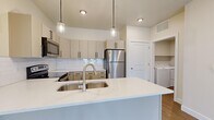 This single-story, one-bedroom, one-bathroom unit offers a thoughtfully laid-out floor plan that maximizes every square foot. The open-concept kitchen and living area serves as the heart of the unit, with the kitchen showcasing cream-toned cabinetry accented by sleek black hardware, a white quartz countertop island with a double stainless steel sink, subway tile backsplash, and a full suite of stainless steel appliances including a refrigerator, range, microwave, and dishwasher. Pendant lighting above the island adds a modern touch. The living area features wood-look flooring and is filled with natural light through large windows and a set of French doors that open to an outdoor view of a landscaped yard.

The bedroom is a comfortable size, finished with plush carpet and a window providing natural light and outdoor views. A dedicated closet provides organized storage.

The bathroom includes a single-sink vanity with quartz countertop, matching cabinetry, a large mirror, and a full shower-tub combo. Wood-look flooring runs throughout the wet areas.

A separate laundry room accommodates a full-size washer and dryer with overhead shelving, adding significant convenience to daily living.
