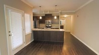 This single-floor space showcases a thoughtfully designed open-concept layout that seamlessly connects the kitchen and living areas. The kitchen serves as the focal point, featuring rich dark wood cabinetry that extends to the upper walls, complemented by light granite countertops that provide an elegant contrast. A peninsula with a built-in sink offers additional prep space and acts as a natural divider between the kitchen and the adjoining living area. Stainless steel appliances, including a refrigerator, electric range, and over-the-range microwave, lend a modern, polished feel to the space. Pendant lighting hangs above the peninsula, adding warmth and focused illumination to the kitchen workspace. Crown molding adds a refined architectural detail throughout, while the rich, dark hardwood flooring flows continuously across the entire floor, grounding the space with a sense of warmth and sophistication. Large shuttered windows invite natural light into the space, brightening the neutral-toned walls. A hallway visible from the main area leads to additional rooms, hinting at a well-organized and functional floor plan. The overall ambiance is clean, contemporary, and well-appointed.