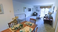 This single-floor space presents an open-concept layout that seamlessly connects the dining and living areas, creating a cohesive and functional environment. The dining area is positioned in the foreground and accommodates a wooden dining table with seating for multiple guests, set with decorative green placemats and accented by floral arrangements that add a touch of warmth.

The transition into the living area is defined by a low-profile metal railing, a subtle yet practical architectural detail. The living room is carpeted throughout, contributing to a comfortable and quiet ambiance. A wall-mounted air conditioning unit provides climate control, while a baseboard heater runs along the perimeter, ensuring year-round comfort.

The living area is furnished with upholstered seating arranged around a central coffee table. Framed artwork is displayed along the walls, adding visual interest to the neutral white walls. A window dressed with fabric valances allows natural light to filter in, offering a view of the surrounding greenery. The overall atmosphere is cozy, functional, and well-maintained, suited for everyday living and casual entertaining.