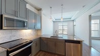 Located at 1719 W Campbell Rd in Garland, Texas, this single-floor residence offers a well-designed layout with contemporary finishes throughout. The kitchen is a standout feature, showcasing grey-toned cabinetry with crown molding detail, granite countertops, a white subway tile backsplash, and a full suite of stainless steel appliances including a range, over-the-range microwave, dishwasher, and refrigerator. Pendant lighting above the granite peninsula adds a polished, functional touch, and the kitchen opens directly to the living area, promoting a connected, open feel.

The living space benefits from a tray ceiling with crown molding detailing and large windows that allow generous natural light and outdoor views. Wood-look flooring runs throughout the main living areas, lending warmth and cohesion to the space. A built-in desk nook near the kitchen provides a practical workspace.

The bedroom is carpeted for comfort and features a ceiling fan and a sizable window. The bathroom includes a glass-enclosed shower, tile flooring, and direct access to a walk-in closet with built-in shelving, adding meaningful storage functionality. Overall, the space combines modern design elements with practical, everyday livability.
