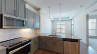 Located at 1719 W Campbell Rd in Garland, Texas, this single-floor residence offers a well-designed layout with contemporary finishes throughout. The kitchen is a standout feature, showcasing grey-toned cabinetry with crown molding detail, granite countertops, a white subway tile backsplash, and a full suite of stainless steel appliances including a range, over-the-range microwave, dishwasher, and refrigerator. Pendant lighting above the granite peninsula adds a polished, functional touch, and the kitchen opens directly to the living area, promoting a connected, open feel.

The living space benefits from a tray ceiling with crown molding detailing and large windows that allow generous natural light and outdoor views. Wood-look flooring runs throughout the main living areas, lending warmth and cohesion to the space. A built-in desk nook near the kitchen provides a practical workspace.

The bedroom is carpeted for comfort and features a ceiling fan and a sizable window. The bathroom includes a glass-enclosed shower, tile flooring, and direct access to a walk-in closet with built-in shelving, adding meaningful storage functionality. Overall, the space combines modern design elements with practical, everyday livability.