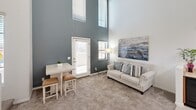 This two-story space is defined by its dramatic double-height ceilings in the main living area, which create a remarkably open and airy atmosphere despite the overall compact footprint. The ground floor hosts the kitchen and living room, where the soaring walls are accented by a bold slate-blue feature wall and multiple windows at varying heights, flooding the space with natural light. A glazed exterior door provides direct outdoor access. The kitchen features warm-toned wood cabinetry, light countertops, black appliances including a range, dishwasher, and refrigerator, and light wood-style flooring, flowing seamlessly into the carpeted living area. A small dining table positioned near the entry door completes this versatile ground-floor layout. A staircase connects both levels, leading to the upper floor, which contains two dedicated office rooms and a full bathroom. Each office is carpeted and benefits from natural light through windows, with one room featuring a ceiling fan. The bathroom is clean and functional, finished with light tile flooring, a pedestal sink, and a shower-tub combination. Throughout, the space maintains a bright, neutral palette with white walls and trim.