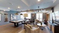 This single-floor space is designed as a multi-purpose game and lounge area, blending recreational amenities with comfortable gathering spaces. The room is anchored by a full-size pool table finished in a rustic wood tone, positioned beneath a pair of industrial-style pendant lights that add a warm, focused glow to the area. Adjacent to the pool table, a shuffleboard table extends the entertainment options available in the space.

The lounge area is thoughtfully arranged with a tufted sofa, upholstered armchairs, and a round wood-topped coffee table, creating an inviting seating cluster suited for conversation or relaxation. A striking sputnik-style chandelier with globe bulbs serves as a visual centerpiece overhead, complementing the layered lighting scheme throughout.

The interior features wood-look flooring, a coffered ceiling with a two-tone blue and white finish, and muted gray-blue walls that contribute to a contemporary, relaxed atmosphere. Large windows along the back wall allow natural light to fill the space and provide views of an outdoor balcony area. Wall art and decorative accents, including a large analog clock, add personality and a lived-in character to the overall design.