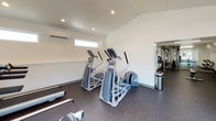 This single-level fitness center is designed to support a comprehensive workout experience within a clean, functional layout. The space is divided into distinct workout zones, each purpose-built for different types of exercise. The main cardio area is bathed in natural light from a series of large windows that line the walls, offering views of the surrounding outdoors and creating an open, airy atmosphere. Recessed ceiling lighting throughout ensures consistent illumination across all areas. The flooring throughout the entire facility consists of durable speckled rubber, providing comfort and safety for all types of exercise.

The cardio zone is outfitted with a rowing machine, stationary bike, elliptical trainers, and a treadmill, catering to a variety of aerobic training preferences. An adjacent strength training area houses a multi-station cable machine with posted instructional workout guides mounted on the wall for user reference. A dedicated free weight section features a full dumbbell rack with a flat weight bench, suitable for a wide range of resistance exercises. A split-unit air conditioning system maintains a comfortable environment year-round. Linear LED lighting in select areas provides bright, even illumination. The crisp white walls and well-organized layout contribute to an efficient and motivating training environment.