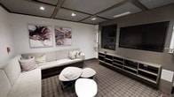 This single-floor space is designed as a contemporary lounge or sitting area, thoughtfully arranged to maximize both comfort and functionality. The room features a light grey upholstered sofa accented with decorative throw pillows, positioned to face a large flat-screen television mounted within a sleek, built-in media unit finished in warm grey tones. The media console offers open shelving below, providing both storage and display space.

A set of modern, organic-shaped white coffee tables are clustered in the center of the room, adding a soft contrast against the patterned grey carpet that grounds the space. The flooring features a subtle geometric design, adding texture without overwhelming the overall neutral palette.

Overhead, a coffered ceiling with recessed lighting provides a structured, architectural element while ensuring even illumination throughout the room. The walls are kept clean and minimal, adorned with two large abstract artworks in muted purples and golds, introducing a refined artistic touch. The overall ambiance is polished and understated, combining modern furnishings with a calm, neutral color scheme suited for relaxation or casual gatherings.