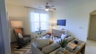 This single-floor space offers a welcoming and thoughtfully designed living area that balances comfort with modern style. The living room serves as the heart of the space, featuring neutral walls that create a calm, airy atmosphere complemented by an abundance of natural light streaming through a large window dressed with sheer white curtains and horizontal blinds. A ceiling fan with an integrated light fixture adds both functionality and a classic touch to the room. The flooring features warm hardwood-style planks, grounded by a chevron-patterned area rug that adds visual interest and texture. Seating is arranged invitingly around a sleek coffee table, with plush gray sofas and an accent armchair creating an ideal gathering area. A wall-mounted flat-screen television sits above a rustic, multi-drawer wooden dresser, which doubles as functional storage and a display surface. Decorative accents, including potted plants, ceramic vases, and stacked books, add personality and a sense of warmth throughout the room. The overall ambiance is light, relaxed, and contemporary, making the space feel both livable and stylish.