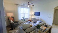 This single-floor space offers a welcoming and thoughtfully designed living area that balances comfort with modern style. The living room serves as the heart of the space, featuring neutral walls that create a calm, airy atmosphere complemented by an abundance of natural light streaming through a large window dressed with sheer white curtains and horizontal blinds. A ceiling fan with an integrated light fixture adds both functionality and a classic touch to the room. The flooring features warm hardwood-style planks, grounded by a chevron-patterned area rug that adds visual interest and texture. Seating is arranged invitingly around a sleek coffee table, with plush gray sofas and an accent armchair creating an ideal gathering area. A wall-mounted flat-screen television sits above a rustic, multi-drawer wooden dresser, which doubles as functional storage and a display surface. Decorative accents, including potted plants, ceramic vases, and stacked books, add personality and a sense of warmth throughout the room. The overall ambiance is light, relaxed, and contemporary, making the space feel both livable and stylish.