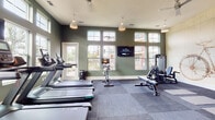 Located at 10952 Netherly Dr in Charlotte, NC, this fitness center is thoughtfully designed to support a wide range of workout routines. The space features sage-green walls accented by crisp white trim, durable modular flooring, and elevated ceilings that contribute to an open, airy atmosphere. Large multi-pane windows line the walls, flooding the interior with abundant natural light and offering views of the surrounding outdoor environment.

The cardio zone is well-appointed with a row of treadmills and stationary cycling bikes, complemented by a wall-mounted flat-screen television for entertainment during workouts. An elliptical trainer and rowing machine provide additional low-impact cardio options. The strength training area includes a multi-functional cable machine, a weight bench with attachments, and a dedicated rack stocked with kettlebells, stability balls, and medicine balls in various sizes. Yoga mats laid out on the floor designate a functional stretching and floor exercise area.

A standout decorative feature is a large artistic bicycle blueprint mural, adding visual character to the strength training corner. Pendant lighting fixtures hang from the ceiling throughout, enhancing both function and aesthetics. An exit door provides direct outdoor access, further connecting the space to its surrounding environment.