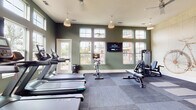 Located at 10952 Netherly Dr in Charlotte, NC, this fitness center is thoughtfully designed to support a wide range of workout routines. The space features sage-green walls accented by crisp white trim, durable modular flooring, and elevated ceilings that contribute to an open, airy atmosphere. Large multi-pane windows line the walls, flooding the interior with abundant natural light and offering views of the surrounding outdoor environment.

The cardio zone is well-appointed with a row of treadmills and stationary cycling bikes, complemented by a wall-mounted flat-screen television for entertainment during workouts. An elliptical trainer and rowing machine provide additional low-impact cardio options. The strength training area includes a multi-functional cable machine, a weight bench with attachments, and a dedicated rack stocked with kettlebells, stability balls, and medicine balls in various sizes. Yoga mats laid out on the floor designate a functional stretching and floor exercise area.

A standout decorative feature is a large artistic bicycle blueprint mural, adding visual character to the strength training corner. Pendant lighting fixtures hang from the ceiling throughout, enhancing both function and aesthetics. An exit door provides direct outdoor access, further connecting the space to its surrounding environment.
