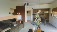 This single-story residence showcases an open-concept layout that seamlessly connects the living, dining, and utility areas. The living room is anchored by a plush olive-green sofa dressed with decorative pillows, positioned on light-toned carpeting that extends throughout the space. A standout design feature is the rich chocolate-brown accent wall, which adds depth and warmth to the interior, complementing the neutral white walls and ceiling. Decorative items, including sculptural figures and greenery, are displayed atop the accent wall's raised ledge, adding visual interest at height. A pendant light fixture with a warm gold tone hangs above the dining area, where a dark-finished table and chairs create a defined eating space, complete with floating wall shelves for added storage or display. Glimpsed through a dedicated alcove, an in-unit washer and dryer provide added convenience. A decorative floral arrangement and candle centerpiece on the coffee table enhance the curated, welcoming ambiance. The overall aesthetic blends earthy tones with warm metallic accents, creating a cohesive and inviting interior atmosphere throughout.