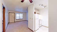 This single-floor space presents a straightforward, functional layout that connects the living area and kitchen in an open, efficient design. The carpeted living room is illuminated by natural light from a large window overlooking what appears to be a private balcony, accessible through a sliding or standard door. A ceiling fan with a light fixture is centrally mounted, adding comfort and airflow to the space. The textured ceiling throughout lends a classic residential feel.

The kitchen is separated from the living area by a partial wall divider, creating a defined cooking space while maintaining an open connection to the rest of the unit. White cabinetry lines the upper walls, providing ample storage, and the kitchen is equipped with a white refrigerator and a white electric range. Track lighting is installed above the kitchen workspace, ensuring well-lit food preparation. The kitchen flooring transitions to light-toned tile, contrasting neatly with the carpeted living area. A thermostat is conveniently positioned on the shared wall between the two spaces. Overall, the unit offers a clean, no-frills interior suited for comfortable everyday living.