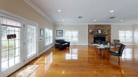 Located at 113 Cherry Court Dr in Greenville, NC, this single-floor space offers a generous, open layout defined by beautifully polished hardwood floors that reflect natural light throughout. The main gathering area is expansive and well-lit, benefiting from multiple large windows fitted with white blinds that allow for flexible light control while framing views of the surrounding exterior. A striking floor-to-ceiling brick fireplace serves as a commanding focal point, adding warmth and character to the space. Crown molding runs along the perimeter of the ceilings, lending a refined, finished aesthetic throughout. Recessed lighting supplements the natural light, ensuring the interior remains bright and inviting. A set of French doors provides a seamless transition to the outdoors, enhancing the sense of openness. Toward the front of the space, a reception or entry area features a pass-through window, a bulletin board, and a counter station, suggesting practical functionality for administrative or community use. The open floor plan allows for versatile furniture arrangements and accommodates multiple seating zones with ease. Neutral wall tones in warm beige complement the rich hardwood flooring, creating a cohesive and welcoming ambiance throughout this well-appointed space.