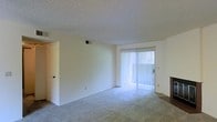 This single-story space features a main living area finished with wall-to-wall carpeting in a neutral tone, contributing to a clean and understated aesthetic. The room is characterized by smooth white walls and a textured popcorn ceiling, which adds a classic residential feel overhead. A prominent sliding glass door fitted with vertical blinds serves as the focal point of the living area, allowing natural light to filter into the space while providing access to an outdoor area. The blinds offer adjustable privacy and light control. An HVAC vent is positioned on the wall near the ceiling, indicating a central climate control system integrated into the unit. A standard interior door is visible along the adjacent wall, suggesting access to additional rooms or storage within the unit. Electrical outlets are distributed at standard intervals along the baseboards. The overall ambiance is bright, open, and move-in ready, presenting a blank canvas suitable for a variety of furnishing arrangements and personal styles.