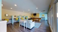 This single-floor amenity space is thoughtfully designed to serve multiple functions within one open, airy layout. The interior is finished with warm wood-look flooring and smooth white ceilings fitted with recessed lighting, creating a clean and inviting atmosphere throughout.

The lounge area anchors the center of the space, featuring a plush light-gray sofa paired with a decorative white console table and a patterned area rug. Cheerful yellow and teal accents are woven throughout the décor, lending the room a vibrant yet cohesive aesthetic.

Along one wall, a dedicated co-working zone offers a row of private workstation cubicles, each equipped with a desk surface and a bright yellow rolling chair — ideal for focused individual work or study.

Toward the back, a full-size billiards table serves as the recreational centerpiece of the space, accompanied by a wall-mounted flat-screen television, making it well-suited for both casual play and entertainment.

A bar-height counter with seating is positioned in the corner, providing a social gathering spot. Complementary yellow decorative elements and framed wall art tie the entire space together, resulting in a lively, multifunctional environment designed for both productivity and leisure.