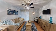 Located in the Elms Plantation community of Charleston, South Carolina, this single-story residence combines comfortable living with a light, coastal-inspired design. The living area serves as the heart of the home, featuring warm hardwood-style flooring, soft blue-toned walls, and a ceiling fan that enhances air circulation and adds a classic touch overhead. A sliding glass door dressed with vertical blinds allows natural light to filter in while providing access to the outdoors. The space is thoughtfully arranged to accommodate both relaxation and entertaining, with room for a full sofa, accent seating, and a central coffee table anchored by a patterned area rug. A wooden sideboard along one wall offers both storage and display space, complementing the warm, natural tones found throughout. Decorative accents in teal and blue echo the coastal character of the greater Charleston region, creating a cohesive and inviting ambiance. The single-floor layout ensures easy accessibility and a seamless flow between living spaces. Overall, the home presents a welcoming atmosphere with practical features suited for comfortable everyday living in one of Charleston's established residential neighborhoods.