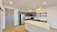 This contemporary single-floor residence showcases a thoughtfully designed open-concept layout with clean lines and modern finishes. The kitchen serves as a central highlight, featuring dark espresso cabinetry, a classic white subway tile backsplash, stainless steel appliances including a full-size refrigerator, range, microwave, and dishwasher, along with a large center island topped with a light quartz countertop. Stylish dome pendant lights with yellow interiors add a pop of color and warmth above the island. Open shelving provides additional display and storage space. Light wood-style flooring flows throughout the main living areas, complementing the bright, neutral color palette. The bedrooms offer plush carpeting and generous natural light through large windows, with one room featuring sliding glass doors that open onto a private balcony overlooking the surrounding development and open landscape. A walk-in closet adds practical storage to the primary bedroom. The bathroom is well-appointed with a rich dark wood vanity, quartz countertop, a large mirror, and a full soaking tub and shower combination. Recessed lighting throughout the space ensures a bright, welcoming ambiance, while the clean, minimalist design creates a versatile and move-in-ready living environment.