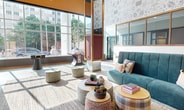 Located at 415 N Lake St in Madison, WI, this 850-square-foot single-floor communal space serves as a dynamic lobby and lounge area designed with a bold, eclectic aesthetic. The space features soaring double-height ceilings that amplify the sense of openness, while expansive floor-to-ceiling windows flood the interior with abundant natural light and offer views of the surrounding streetscape. The main lounge area is anchored by a tufted teal sectional sofa paired with an assortment of upholstered ottomans and low-profile coffee tables, creating an inviting social hub. Decorative elements include a suspended gold-framed plant shelf with trailing greenery, a large abstract blue wall art piece, and eye-catching botanical-patterned wallcoverings. Polished concrete flooring transitions to layered area rugs, adding texture and warmth throughout. A dedicated entertainment zone features a foosball table positioned near the windowed facade. Adjacent to the lounge, a kitchenette area with stainless steel appliances and a patterned tile floor provides practical amenities. A small desk nook with curated wall art offers a quiet workspace. The overall ambiance balances modern industrial elements—exposed ductwork, metal accents—with warm wood paneling and vibrant pops of pink and orange, resulting in a lively yet comfortable communal environment.