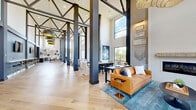 Located at 300 Highway 81 in McDonough, Georgia, this expansive single-story space makes an immediate architectural statement with its soaring double-height ceilings and bold exposed steel truss framework overhead. Light-toned hardwood flooring runs throughout, anchoring the open, flowing layout that connects multiple distinct functional zones seamlessly.

The main lounge area features two cognac leather sofas flanking a sleek linear fireplace set into a floor-to-ceiling shiplap accent wall, framed by curated artwork and decorative sconces. Nearby, a secondary seating arrangement with linen-upholstered chairs and a round coffee table faces oversized floor-to-ceiling windows that flood the space with natural light and offer views of the surrounding landscape.

A generously sized dining area with a dark table and upholstered seating anchors the central zone, while a built-in bar and kitchenette features navy cabinetry with brass hardware, dark herringbone tile backsplash, stainless appliances, and a dramatic quartz-topped island accented with bold blue chairs. Flat-screen displays are mounted above the bar cabinetry. A counter-height workspace with bar seating lines one wall, ideal for casual work or socializing. Statement black-and-gold pendant lights and a sculptural chandelier complete the refined yet welcoming ambiance throughout.