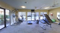This fitness center offers a dedicated space for exercise and wellness, thoughtfully designed to support a range of cardio-focused workouts. The room is outfitted with multiple treadmills, an elliptical trainer, and additional cardio equipment, providing ample options for residents or members to maintain an active lifestyle. The flooring features durable, low-pile carpet suitable for high-traffic exercise use, while the warm yellow walls contribute to an energizing yet welcoming atmosphere. Overhead, a ceiling fan and recessed lighting ensure the space remains comfortable and well-lit throughout the day. A wall-mounted flat-screen television adds convenience for entertainment during workouts. Large sliding glass doors and wide windows line the far wall, flooding the room with natural light and offering clear views of the outdoor surroundings, creating an open and airy feel despite the enclosed setting. The single-floor layout keeps everything easily accessible, and the open floor plan allows for comfortable movement between equipment. Ventilation is supported by both the ceiling fan and visible HVAC ductwork, ensuring a comfortable environment during intense exercise sessions. Overall, this fitness center is a practical and inviting space designed to meet everyday workout needs.