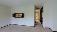 Located at 1942 Mackenzie Dr in Columbus, Ohio, this multi-level home spans three floors, offering a well-distributed layout across its levels. The main living area is finished with wall-to-wall carpeting and neutral white walls, creating a clean and open atmosphere. A notable architectural detail is the built-in pass-through window with dark wood shutters connecting the living room to the kitchen — a classic design element that promotes an open sightline between the two spaces. The kitchen, visible through the pass-through, features light wood cabinetry and standard appliances. A hallway leads from the living area toward additional rooms, with a multi-pane door providing a natural transition point within the home. A dark wood staircase with open baluster railing is prominently situated near the hallway, connecting the main level to the upper and lower floors. The staircase's dark wood tones serve as a strong visual contrast against the lighter walls and carpeting throughout. The home's straightforward layout and classic interior details offer a practical and functional living environment with room to personalize across all three floors.