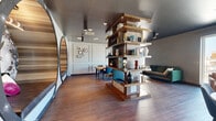 Located at 247 N Fourth Ave in Tucson, Arizona, this thoughtfully designed single-floor office space offers a compelling blend of creative architecture and professional functionality across two distinct office areas. Rich hardwood flooring runs throughout, grounding the space with warmth against the dark, high-gloss ceiling that adds a sleek, modern contrast.

A freestanding, open-concept wood shelving tower serves as a central dividing element, elegantly separating the collaborative lounge area from the conference space without enclosing either. The conference office features classic wainscoting wall paneling adorned with framed artwork, paired with a solid wood conference table and teal upholstered chairs — conveying a polished yet inviting atmosphere.

The lounge area offers flexible seating including a sofa and accent chairs, anchored near a large floor-to-ceiling window that floods the space with natural light and frames an open view of the Tucson cityscape. A round table with additional seating provides an informal meeting or breakout zone.

One of the most striking architectural features is a large circular nook framed in dark metal with warm wood slat cladding on the interior, housing a built-in leather bench seat beneath decorative botanical wallpaper. This immersive pod-style retreat offers a private, cozy counterpoint to the open office environment, making the space as imaginative as it is functional.