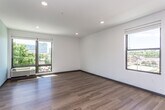 This single-floor unit showcases a clean, contemporary design throughout, characterized by crisp white walls, dark baseboards, and warm hardwood-style flooring that flows seamlessly across all rooms. Recessed lighting enhances the bright, open atmosphere, complemented by multiple windows that invite generous natural light and provide views of the surrounding urban landscape.

The open living area is generously proportioned, offering flexible arrangement possibilities. Large windows frame outdoor views of neighboring buildings and green tree canopies, adding vibrancy to the interior. The kitchen is efficiently designed, featuring stainless steel appliances including a refrigerator, electric range, over-the-range microwave, and dishwasher. Grey cabinetry with sleek hardware pairs with light countertops and a soft blue-green accent wall, creating a modern, functional workspace.

A dedicated laundry area houses a full-sized stacked washer and dryer unit, offering added convenience. The bathroom is well-appointed with a combined shower and soaking tub, a dark grey vanity with ample drawer storage, white countertops, and brushed nickel fixtures. The bedroom provides a comfortable, well-lit retreat with a window and built-in shelving nearby. Overall, the unit presents a cohesive, modern aesthetic suited for practical urban living.