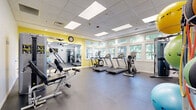 Located at 5707 Luxemburg St in Rockville, MD, this fitness center offers a well-appointed exercise room designed to support a comprehensive workout routine. The space features a drop-panel ceiling with recessed and flush lighting panels that ensure consistent, bright illumination throughout. A row of large windows along one wall floods the room with natural light and provides pleasant views of surrounding greenery, creating an energizing atmosphere for exercise.

The cardio section is equipped with multiple treadmills and an elliptical trainer, all positioned to take advantage of the outdoor views. A multi-station cable strength machine serves as the centerpiece for resistance training, accommodating a variety of exercises. Additional fitness accessories include stability balls, resistance bands, medicine balls, and dumbbells, organized neatly on a dedicated storage rack. A wall-mounted flat-screen television adds entertainment during workouts.

Full-length mirrors on one wall visually expand the space and allow users to monitor their form. The durable rubber flooring provides cushioning and traction throughout. Accent walls in a warm yellow tone add a vibrant, motivating energy to the room, complementing the otherwise clean white walls. A wall clock and sanitation station complete the practical amenities of this fully equipped community fitness center.