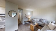 This single-floor residence offers a thoughtfully designed layout that balances comfort and functionality throughout. The living area is warm and inviting, featuring plush carpeting, textured ceilings, and panel-accented walls that lend a distinctive character to the space. Large windows allow natural light to fill the room, creating an airy atmosphere, while a wall sconce and decorative artwork add a cozy, curated touch.

The kitchen is efficiently arranged in an L-shaped configuration, featuring white cabinetry with brushed nickel hardware, dark countertops, and wood-look vinyl flooring. White appliances, including a range, over-the-range microwave, and refrigerator, are seamlessly integrated into the design, offering everything needed for everyday cooking.

The bedroom is a serene retreat, complete with carpeted flooring, a closet with built-in shelving, a window with natural light, and panel-style walls that echo the design language found throughout the home. An additional room visible in the floor plan provides flexible space for various uses.

The bathroom features retro-style mosaic tile flooring and light blue ceramic wall tiles, giving it a charming vintage character. Built-in shelving provides practical storage alongside a pedestal sink. Overall, this residence presents a cohesive, comfortable living environment with a neutral palette and functional design throughout.