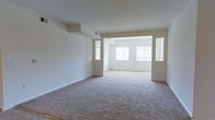 This single-floor space offers an open, airy layout defined by clean lines and a neutral color palette throughout. The main living area features wall-to-wall plush carpeting in a warm beige tone, providing a comfortable foundation underfoot. Smooth white walls and white ceilings contribute to a bright, expansive feel, while HVAC vents are thoughtfully integrated into the ceiling for a clean aesthetic.

A wide, open passageway connects the primary living area to an adjacent room, creating a seamless flow between spaces. The connected room is bathed in natural light, courtesy of multiple large windows — including a prominent trio of windows along the far wall and additional narrow sidelight windows flanking the passage — allowing daylight to penetrate deep into the interior.

The recessed ceiling detail at the passageway adds a subtle architectural element, lending visual interest without disrupting the overall minimalist character of the space. The consistent use of light tones across walls, ceilings, and flooring creates a cohesive, welcoming ambiance that feels open and versatile, well-suited to a variety of furnishing and layout configurations.