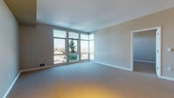 This single-floor unit is thoughtfully designed with an open-concept layout that connects the living, dining, and kitchen areas into one cohesive space. The kitchen is a standout feature, finished with bold yellow cabinetry, a dark countertop, a stainless steel sink, and white appliances including a range, microwave, and refrigerator. Hardwood-style flooring runs throughout the kitchen and laundry areas, while soft neutral carpet covers the living and sleeping spaces.

The spacious living area is defined by large corner windows that flood the room with natural light and offer expansive views of the surrounding neighborhood and beyond, including glimpses of water and greenery. A half-wall separates the kitchen from the living area, and track lighting along the kitchen soffit provides additional illumination.

The bedroom is generously proportioned, also benefiting from wide windows with outdoor views and a baseboard heater for year-round comfort. The bathroom is finished with hardwood-style flooring, a soaking tub with a handheld showerhead, grab bars for accessibility, a wall-mounted sink, and dual mirrors with overhead lighting. A dedicated laundry area houses a full-size front-loading washer and dryer, adding everyday convenience to this well-appointed unit.