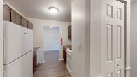 This single-floor unit offers a functional and straightforward layout connecting several distinct living spaces. The galley-style kitchen is equipped with white appliances, including a coil-burner electric range, refrigerator, and dishwasher, complemented by warm-toned wood cabinetry above and below the counters. A textured ceiling and flush-mount lighting illuminate the space, which opens directly into an adjacent living or dining area. The living area features wood-look hard-surface flooring, a baseboard heating system, and a window-mounted air conditioning unit that provides natural light and outdoor views. Bedrooms are finished with neutral carpet and feature windows that bring in additional natural light, along with dedicated closet space equipped with a hanging rod and shelf. The bathroom includes a tub-shower combination with patterned tile surround, a mosaic tile floor with a decorative dot motif, a vanity with a marble-style countertop, and built-in storage. Throughout the unit, white walls and textured ceilings create a clean, neutral backdrop, while baseboard heaters are present in multiple rooms for consistent climate control.