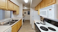 This single-story residence offers a functional and comfortable layout across several well-defined living spaces, all finished with light wood-tone flooring that lends a cohesive, clean aesthetic throughout. The kitchen is designed as a galley-style space, featuring light wood cabinetry, ample upper and lower storage, a double-basin sink, electric range, refrigerator, and built-in microwave. Notably, an in-unit washer is located just off the kitchen, adding everyday convenience. The adjacent dining area accommodates a round table with seating for four, lit by a pendant light fixture, and opens visually toward the kitchen. The living room is bright and open, highlighted by a large sliding glass door that invites natural light and provides outdoor access. Multiple bedrooms are present throughout the unit, each featuring windows with blinds and curtains, closet access, and sufficient space for standard bedroom furnishings. Two full bathrooms serve the residence, each equipped with a single vanity featuring light wood cabinetry, a wall mirror, and a shower-tub combination with a curtain. The overall design is neutral and practical, with white walls and consistent finishes creating a unified, move-in-ready environment.