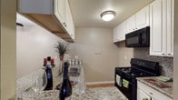 This single-floor residence offers a thoughtfully designed layout centered around a functional and stylish kitchen. The kitchen is a standout feature, showcasing granite countertops that provide ample prep space and a durable, attractive surface. White upper cabinetry lines the walls, offering generous storage while contributing to a bright, clean aesthetic. Black appliances, including a gas range and overhead microwave, provide a bold contrast against the lighter tones of the cabinetry and counters. A stainless steel sink is neatly integrated into the countertop, adding a practical and modern touch. The kitchen is illuminated by a flush-mount ceiling light that evenly brightens the space. Neutral-toned walls and light hardwood flooring create a warm, cohesive atmosphere throughout. The overall design balances practicality with visual appeal, making the kitchen both a highly functional workspace and an inviting area. The single-floor layout ensures easy accessibility and a seamless flow throughout the residence, making it a comfortable and efficient space for everyday living.