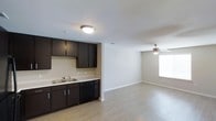 This single-floor residence offers a clean, contemporary layout that seamlessly blends the kitchen and living areas into one open, functional space. The kitchen is outfitted with rich, dark espresso cabinetry featuring sleek bar-pull hardware, light-toned countertops, a double-basin sink, and a black dishwasher — creating a striking contrast against the neutral walls. A flush-mount ceiling light provides focused illumination over the kitchen workspace. The space transitions effortlessly into a spacious living area, where light wood-style laminate flooring extends throughout, lending warmth and visual continuity. The living area benefits from natural light streaming through a window, and a ceiling fan with an integrated light fixture ensures comfortable airflow year-round. Neutral, light gray walls provide a versatile backdrop suitable for a variety of décor styles. Smooth transitions between rooms, consistent flooring, and a cohesive color palette give the space a polished, move-in-ready feel. The open-concept design maximizes the sense of space, making the layout feel both airy and practical for everyday living.