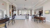 Located at 1000 Worthington Ln in Spring Hill, TN, this single-floor community clubhouse offers a generously sized, open-concept layout designed for both comfort and functionality. The interior is finished with large-format light tile flooring throughout, crown molding, and soaring tray ceilings that enhance the sense of volume and airiness across the entire space.

The central lounge area serves as the heart of the clubhouse, anchored by a dramatic floor-to-ceiling tiled fireplace wall with a sleek linear fireplace and a wall-mounted TV. Comfortable seating arrangements are complemented by a ceiling fan and recessed lighting. Expansive windows along the rear wall flood the space with abundant natural light and provide views of the outdoor pool and surrounding greenery.

Adjacent to the lounge is a casual dining area featuring high-top tables and bar seating, positioned directly beside a bank of large windows and a glass-panel door that opens to the exterior. The kitchen is well-appointed with rich dark-wood cabinetry, granite countertops, stainless steel appliances, a breakfast bar with leather bar stools, and striking blue pendant lights overhead.

A dedicated office or leasing area occupies one section of the space, furnished with wooden desks, guest seating, and access to a back office with built-in cabinetry. The overall ambiance is bright, welcoming, and polished throughout.
