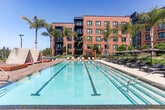 Located at 1200 Rickabaugh Way in Santa Clara, California, this thoughtfully designed outdoor amenity space offers a resort-inspired experience within a modern residential community. The centerpiece is a generous swimming pool framed by tall palm trees, complemented by a separate spa and an expansive sun deck lined with chaise lounges — all enjoying sweeping views of the surrounding Santa Clara landscape under California's abundant sunshine.

Adjacent to the pool area, a covered outdoor kitchen and dining terrace provides a well-equipped barbecue station beneath a slatted pergola, with multiple dining tables and seating for social gatherings. Large glass-paneled doors connect the terrace to the building's interior, allowing seamless indoor-outdoor flow.

Lush, curated landscaping defines the central courtyard, where raised concrete planters overflow with native grasses, flowering plants, and mature trees. A manicured lawn area offers open green space, accented by a warm wood-framed pergola cabana with cushioned lounge seating — ideal for relaxing in a more intimate setting. Whimsical orange bean bag chairs add a playful touch to the lawn.

Throughout the space, high-end materials including large-format tile pavers, warm wood decking, and architectural steel details reflect a contemporary design aesthetic that is both functional and visually refined.