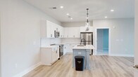Located at 74 Callaway Dr NW in Calabash, NC, this well-designed 2-bedroom, 2-bathroom single-floor home offers a clean, modern aesthetic throughout. The open-concept kitchen and living area serves as the heart of the home, anchored by a central island with a white quartz countertop, undermount sink, and matte black faucet. White shaker cabinetry, a classic subway tile backsplash, and stainless steel appliances—including a refrigerator, range, and microwave—complete the kitchen. Stylish pendant lights with cylindrical glass shades hang above the island, adding a refined touch. Light wood-look flooring runs throughout the main living areas, enhancing the bright, airy feel. The living area features sliding glass doors that open to a private balcony with views of the surrounding community, allowing generous natural light to fill the space. Each bedroom is finished with plush carpet, a ceiling fan, and access to natural light. The spacious primary bedroom includes its own sliding door to a balcony. The bathrooms feature white vanities, framed mirrors, and contemporary brushed nickel hardware. A dedicated walk-in closet with built-in shelving and hanging rods provides excellent storage capacity, rounding out this thoughtfully appointed home.