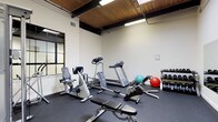 This fitness room offers a thoughtfully organized workout space with a distinctive architectural character. The ceiling features warm wood planking accented by exposed dark beams, creating an industrial-yet-inviting atmosphere, while overhead fluorescent lighting ensures the space is well-lit throughout. White walls and dark rubber flooring provide a clean, functional aesthetic that keeps the focus on the equipment.

The room is outfitted with a well-rounded selection of cardio equipment, including a treadmill, recumbent bike, upright bike, and a rowing machine, offering diverse aerobic training options. A multi-function cable machine serves as a centerpiece for strength training, complemented by an adjustable workout bench. A tiered dumbbell rack holds a comprehensive range of free weights, and exercise balls in multiple sizes round out the functional fitness offerings.

Large wall-mounted mirrors along one side visually expand the space and allow users to monitor their form during workouts. A wall-mounted television adds entertainment during exercise sessions. A grid-paned interior window connects the room to an adjacent space, adding architectural interest and openness. The layout provides ample room to move freely between stations, making this a practical and complete fitness facility.