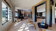 Located at 500 Lama Dr in Summerville, South Carolina, this single-story community clubhouse is a thoughtfully designed gathering space that blends contemporary aesthetics with warm, inviting materials. Light hardwood flooring runs throughout, unifying the various distinct zones within the open-plan layout.

The main social lounge features soaring ceilings, a dramatic dark-brick fireplace surround, and a striking geometric metal-and-wood shelving wall. Seating areas are furnished with mid-century modern chairs and plush sectional sofas, creating an atmosphere ideal for both relaxed socializing and casual gatherings. Large floor-to-ceiling windows and glass doors flood the interior with natural light and offer direct views of the outdoor pool and pergola area.

A fully equipped demonstration kitchen anchors one section of the space, complete with stainless steel appliances, a subway tile backsplash, dark cabinetry, and a generous white-quartz island with bar seating beneath bold pendant lights. Adjacent to the kitchen, a conference-style dining room accommodates a large live-edge wood table surrounded by upholstered chairs, accented by textured wallcovering and curated artwork.

Additional areas include a sunlit casual lounge with a wall-mounted TV, a games zone featuring a shuffleboard table, and multiple intimate seating nooks offering outdoor views. The cohesive design throughout balances bold navy and natural wood tones with an open, light-filled ambiance.